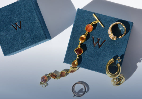 A multi-colored gemstone gold bracelet with gold earrings scattered on top of a white background with two blue jewelry boxes with a gold W on them